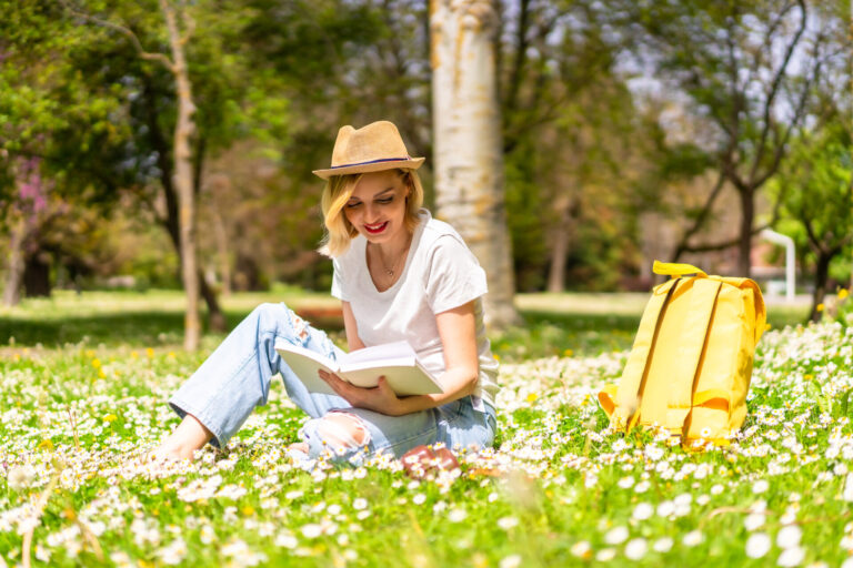 Woman sitting on a grassy field in a park, reading a book with a yellow backpack beside her.