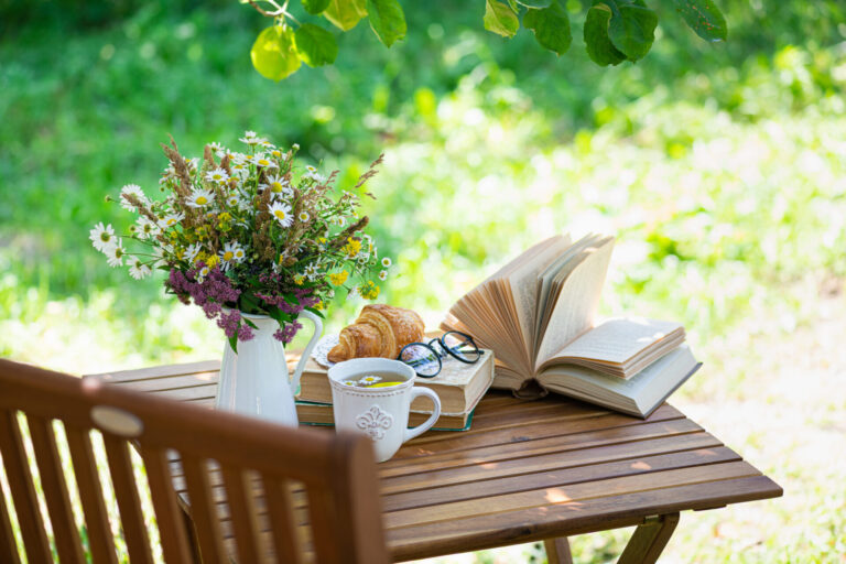 Outdoor wooden table with daisies, an open book, a croissant, tea, and glasses in a sunlit garden.