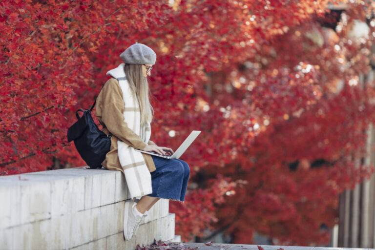 Woman in a gray beret and scarf sits on a concrete ledge, typing on a laptop with a backdrop of red autumn leaves.