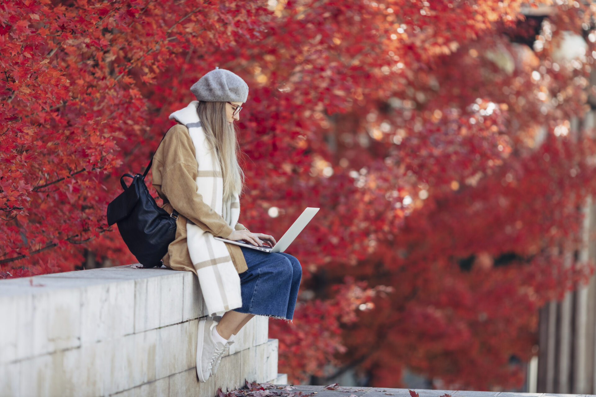 Woman in a gray beret and scarf sits on a concrete ledge, typing on a laptop with a backdrop of red autumn leaves.