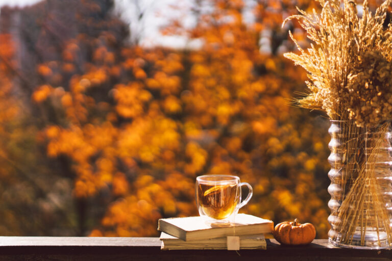 Tea in a glass mug sits on stacked books with a small pumpkin and dried grasses in a vase, autumn orange background.